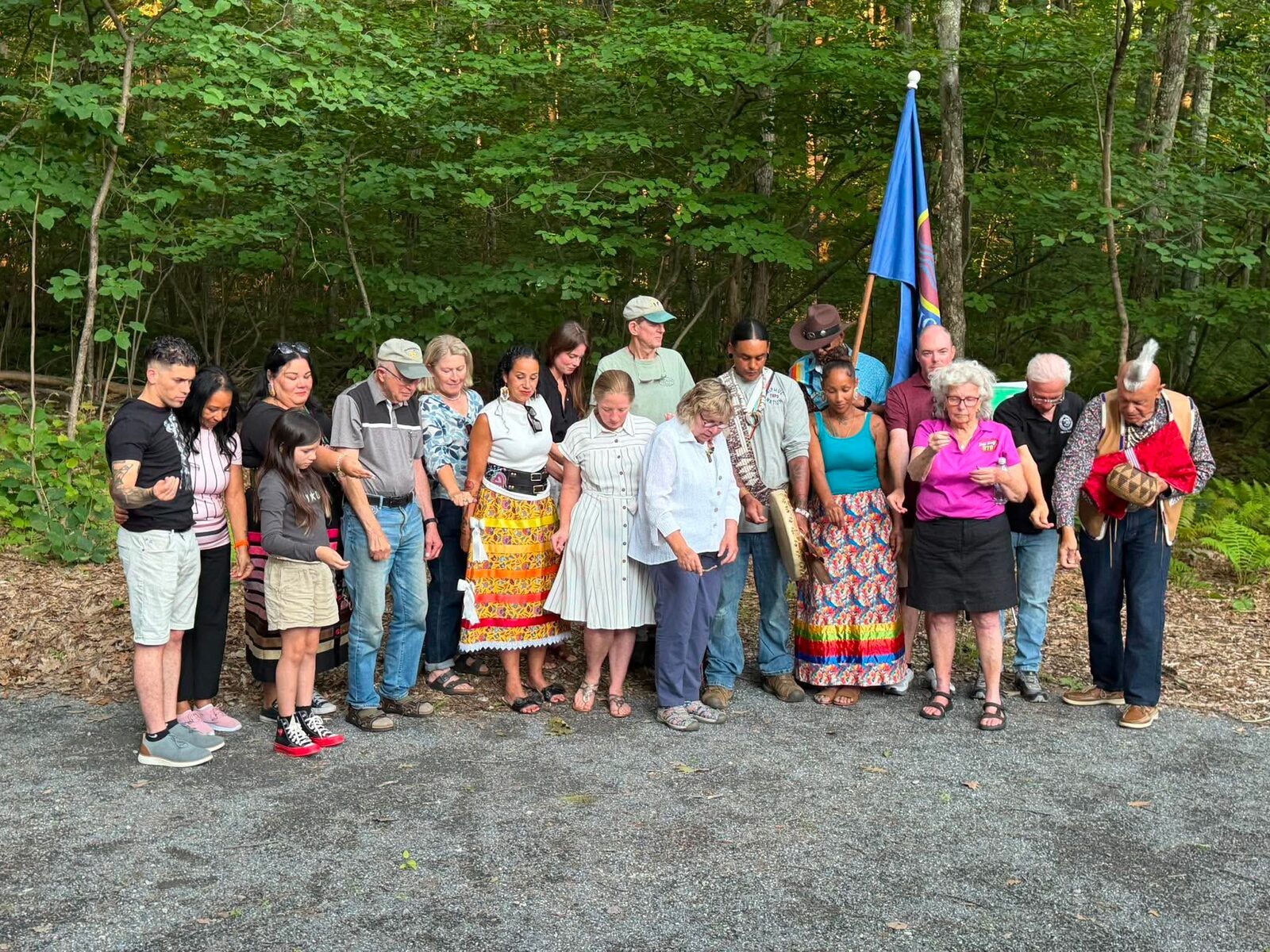 Community members standing hand in hand in a circle around the Nipmuc Nation flag during the bench naming ceremony