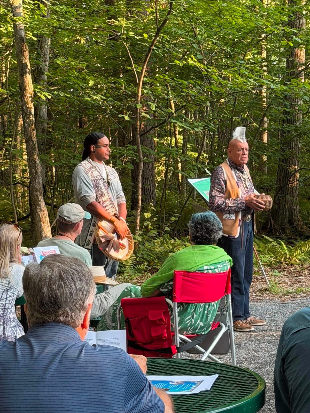 Tribal members speaking and holding a hand drum during the bench naming ceremony in the woods