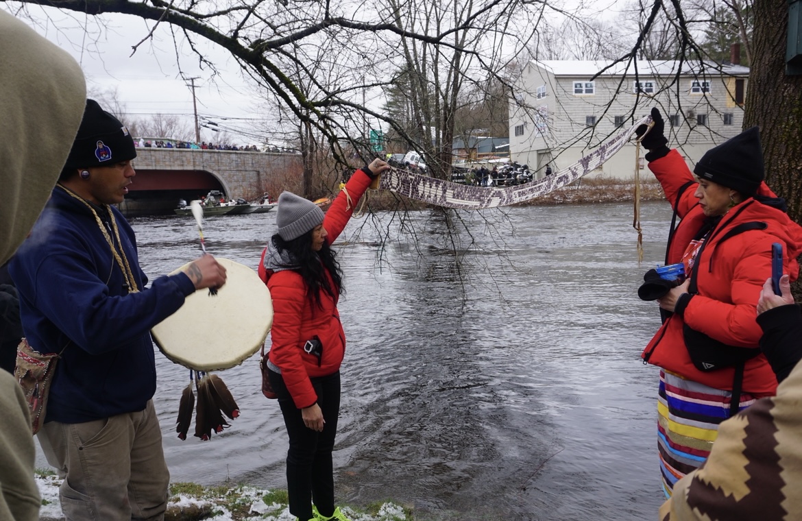 Water ceremony participants holding a woven paddle-shaped banner over the river