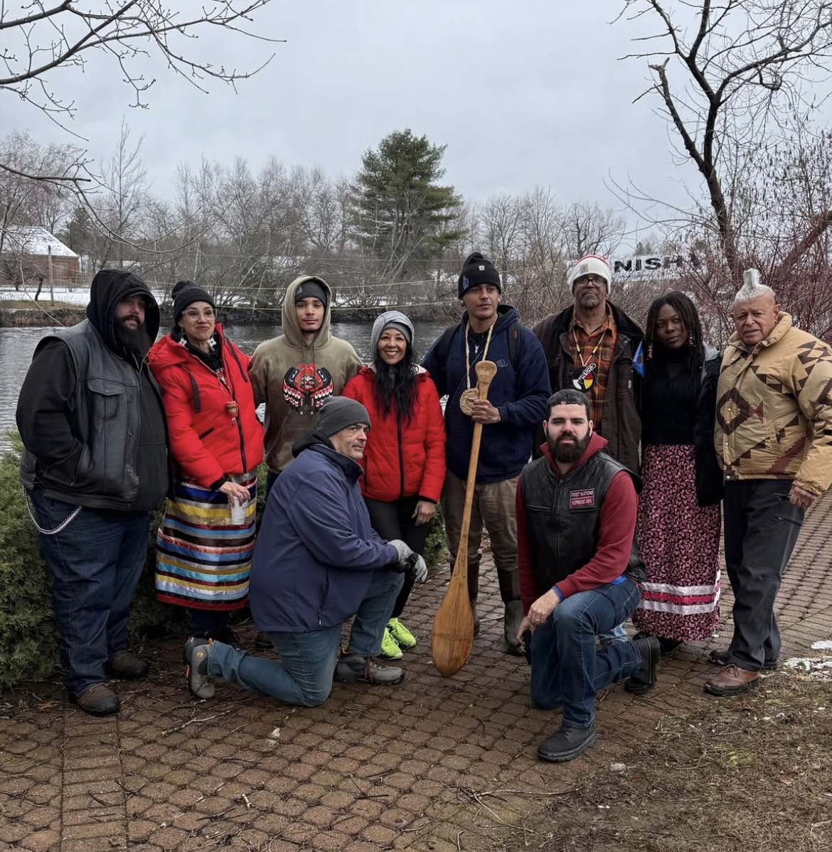 Group portrait beside the river holding a wooden canoe paddle