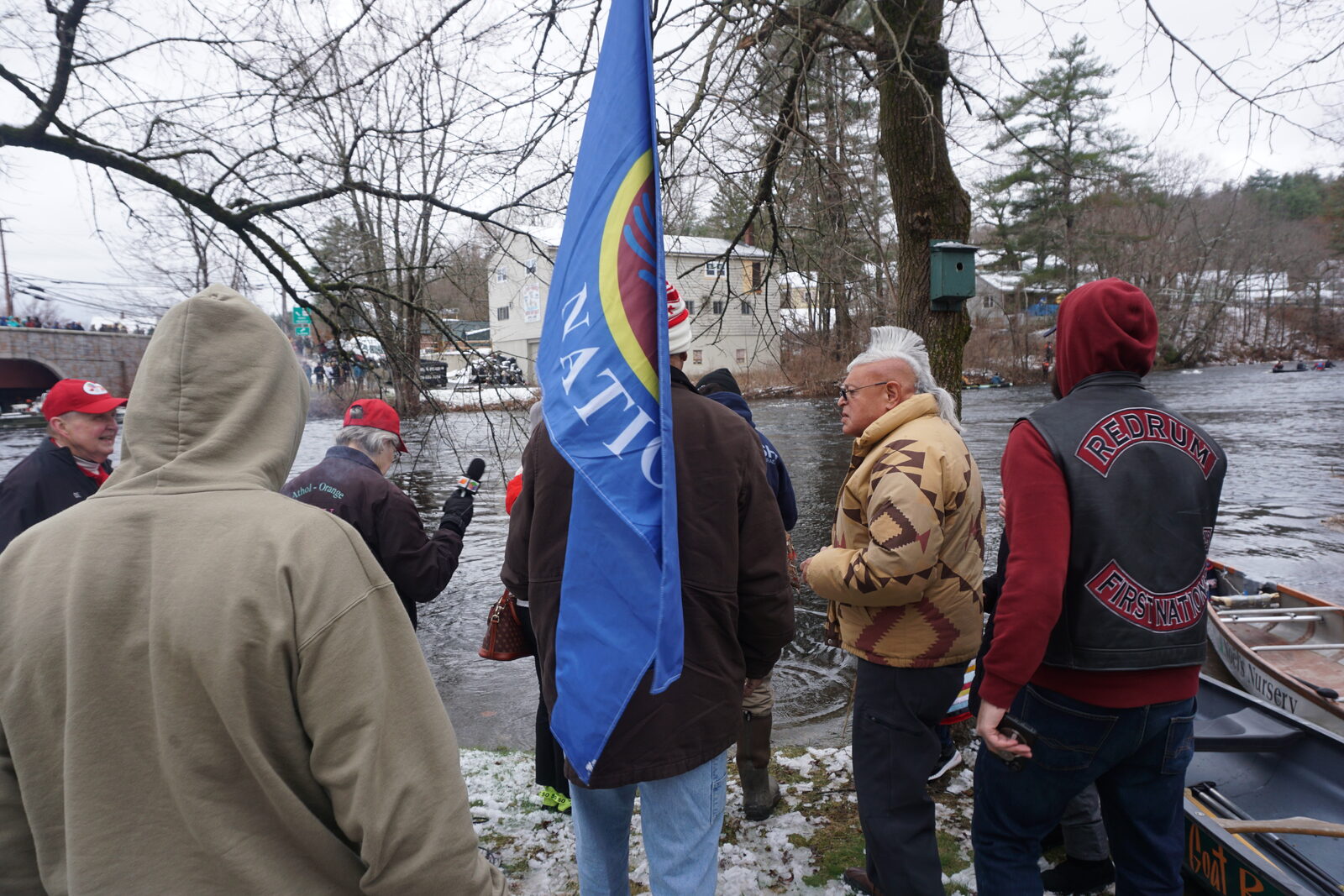 Community members carrying the Nipmuc Nation flag while speaking with a local news reporter by the river