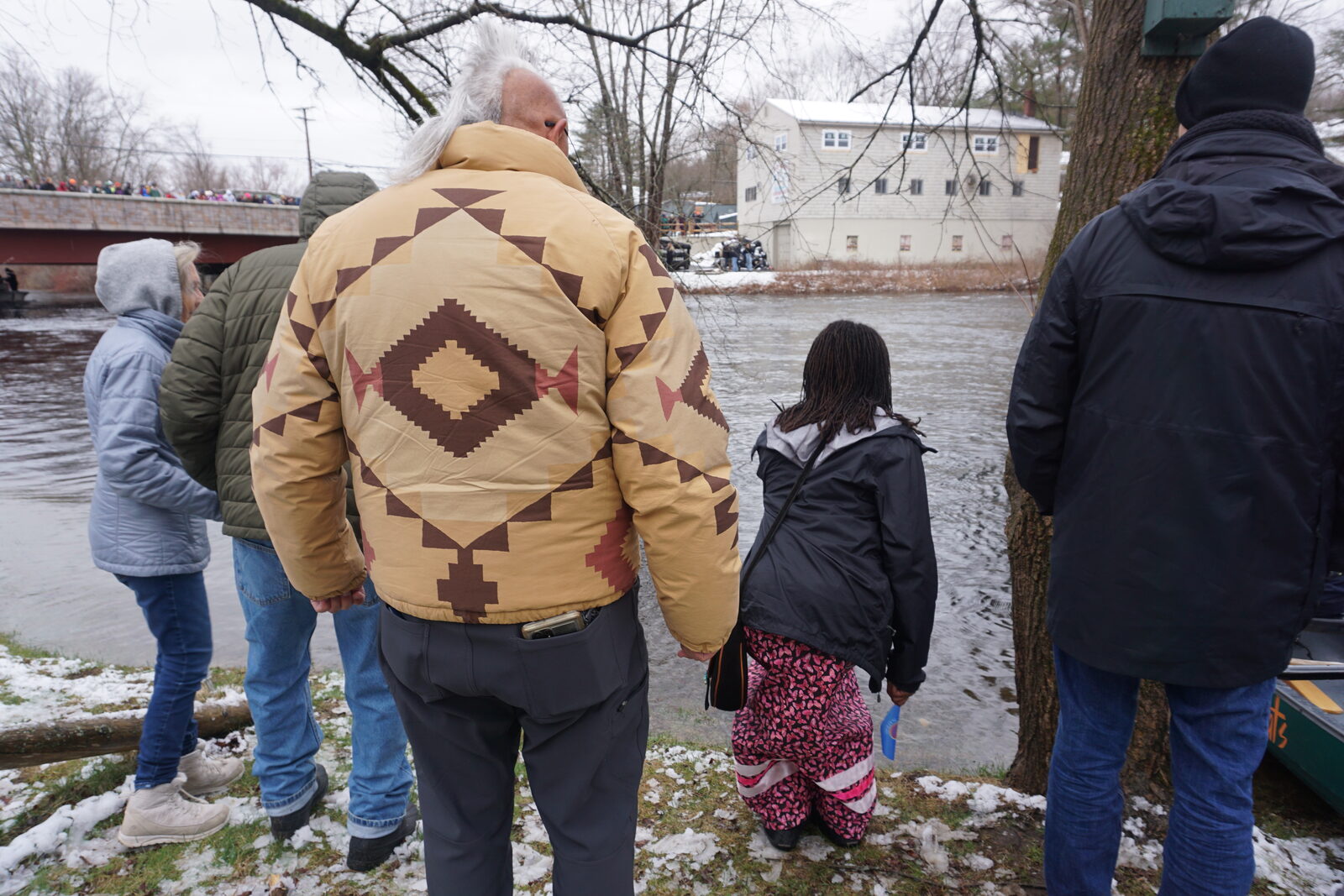 Community members standing hand in hand along the riverbank watching the canoes