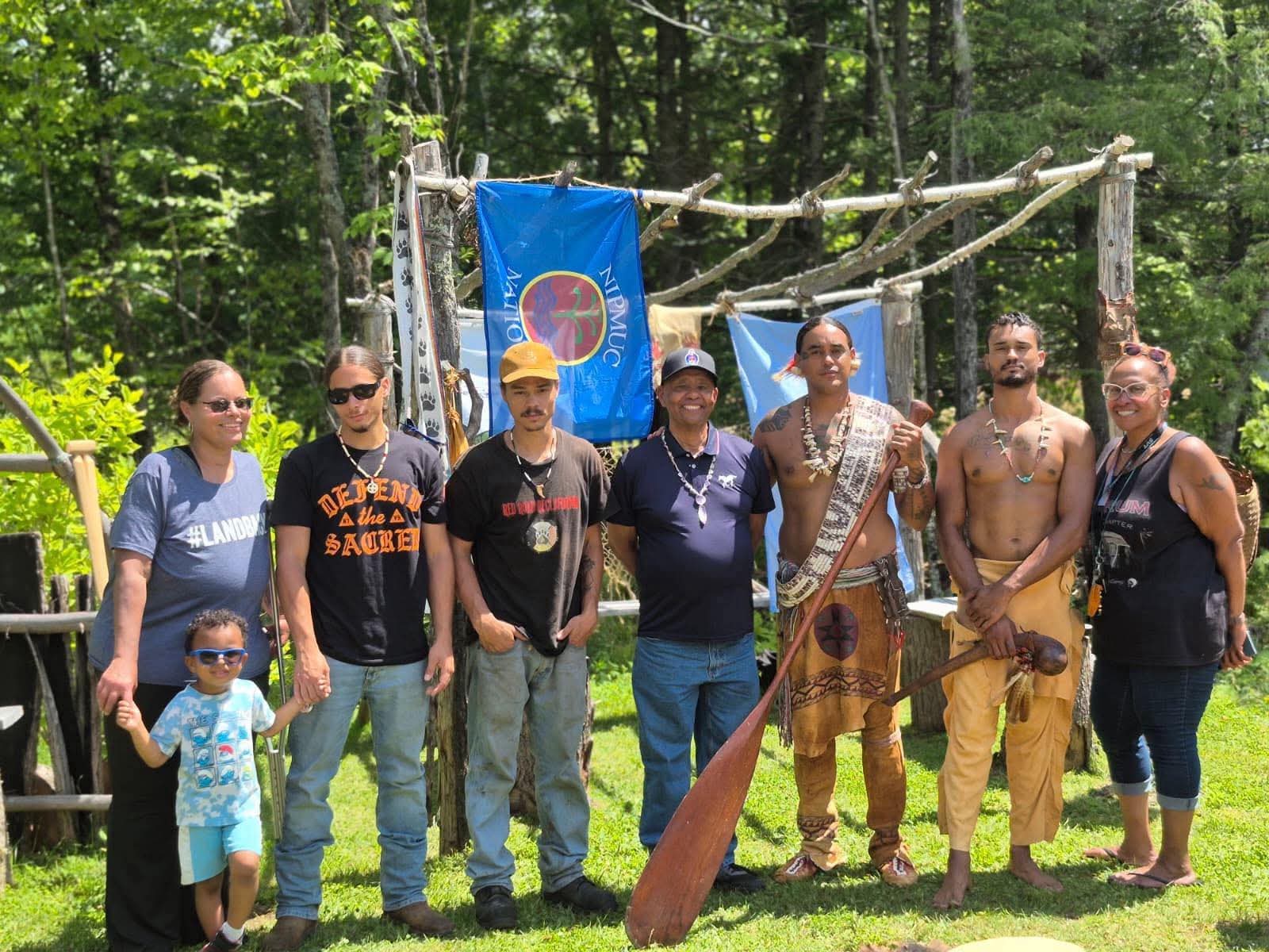 Group photo at a mishoon-building frame with Nipmuc Nation flags