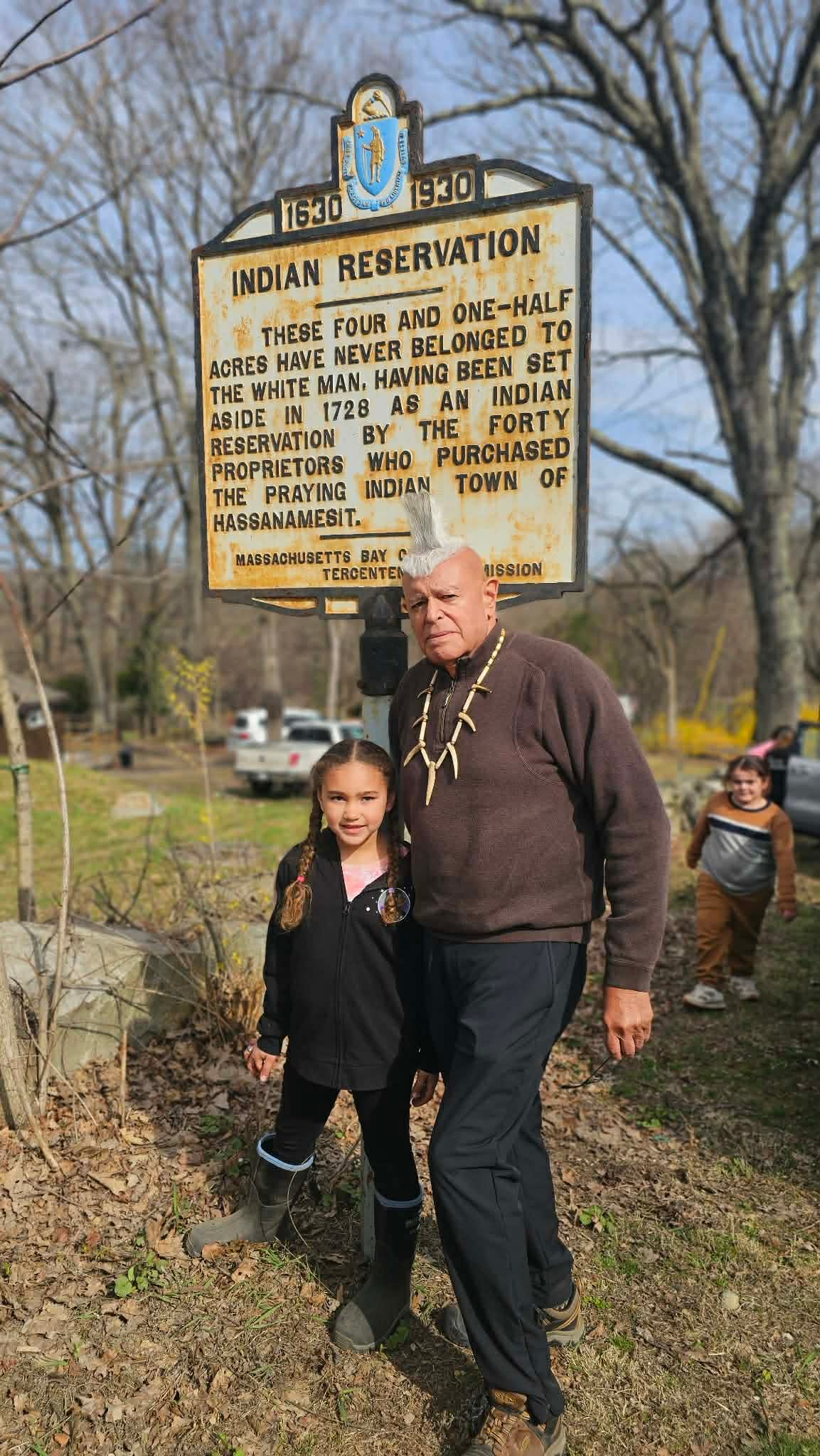 An elder and his granddaughter standing beside the Indian Reservation historical marker at Hassanamesit
