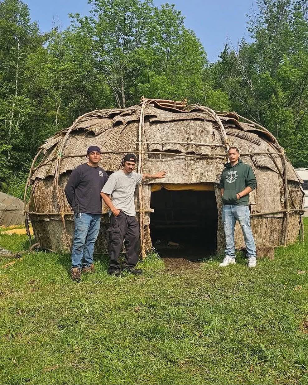 Three men standing in front of a completed bark-covered wigwam