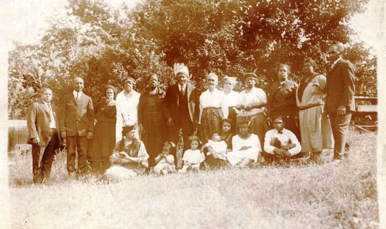 Historical group portrait of an extended family, including a man wearing a feathered headdress, seated and standing outdoors