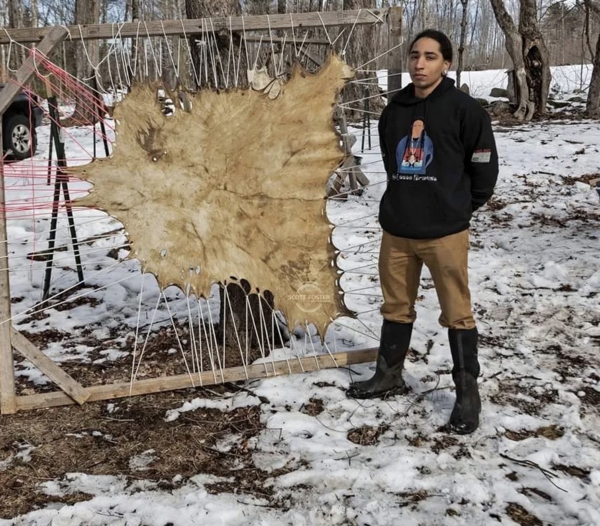 A young man standing beside a deer hide stretched on a wooden tanning frame