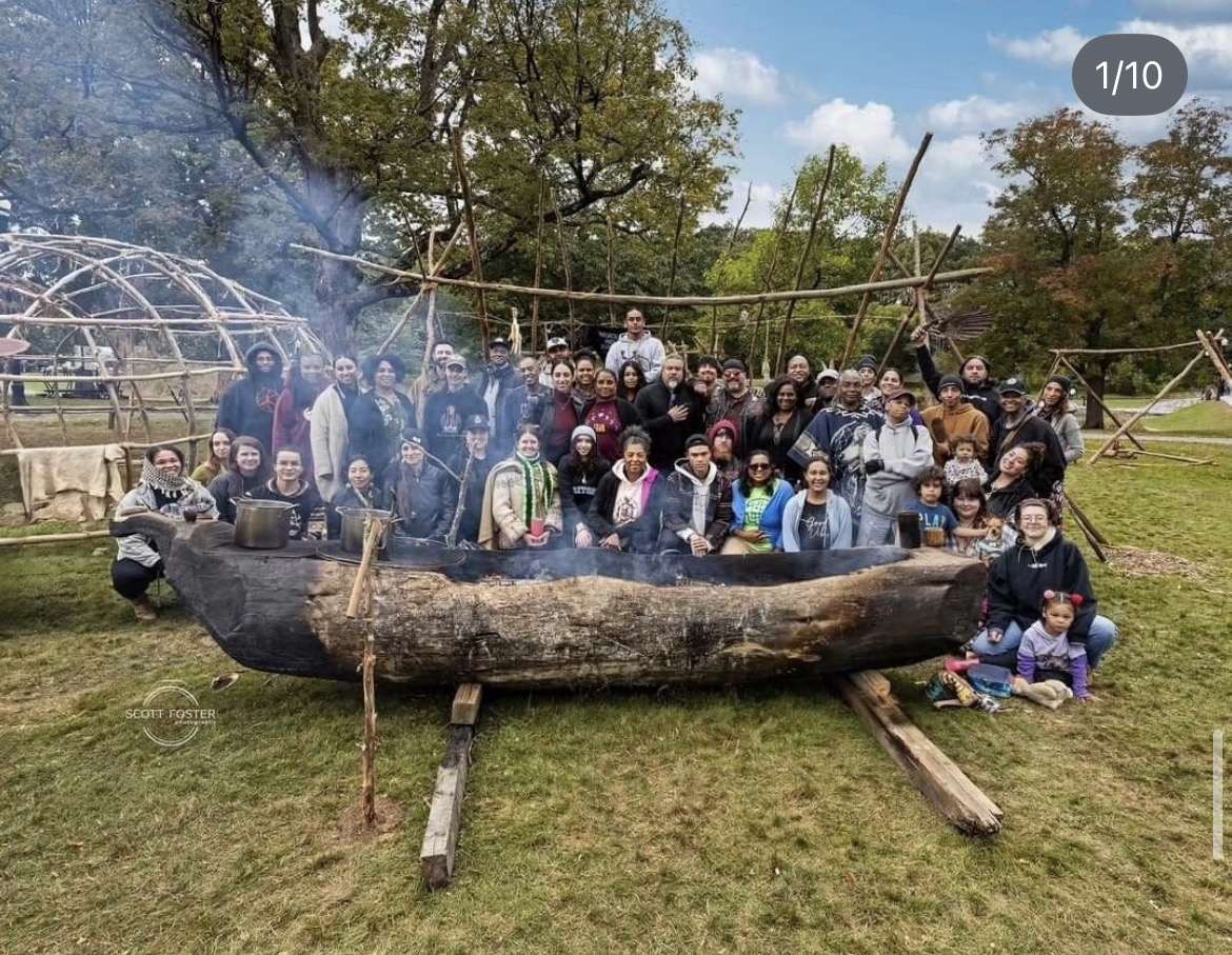 Large group of community members gathered around a carved mishoon (dugout canoe) beside a wigwam frame
