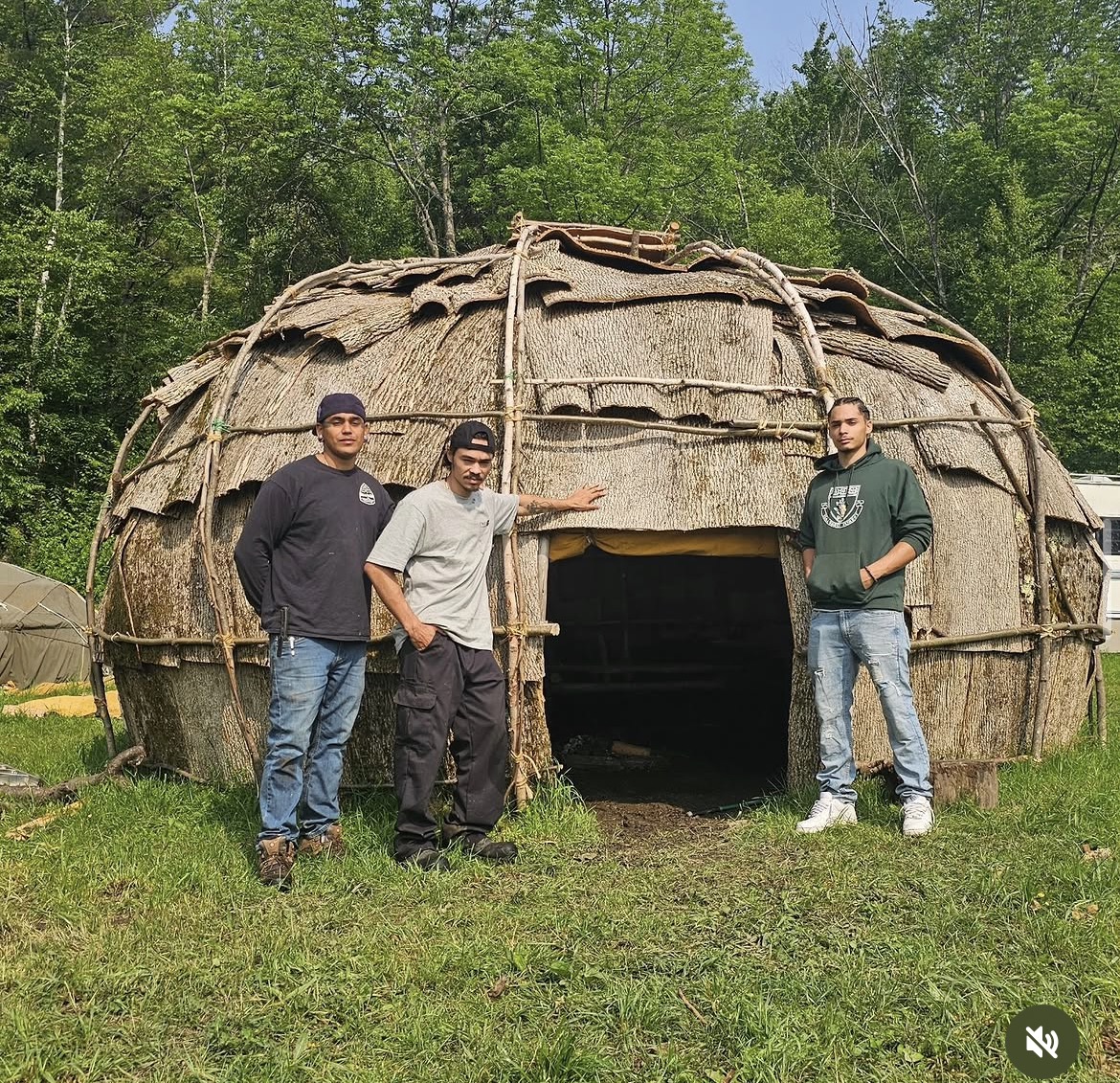 Three community members standing in front of a completed bark-covered wigwam