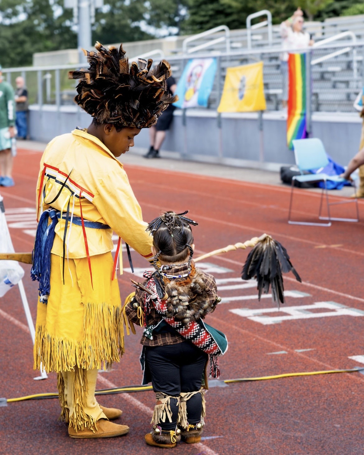 A young dancer in a feathered headdress with a child at the powwow
