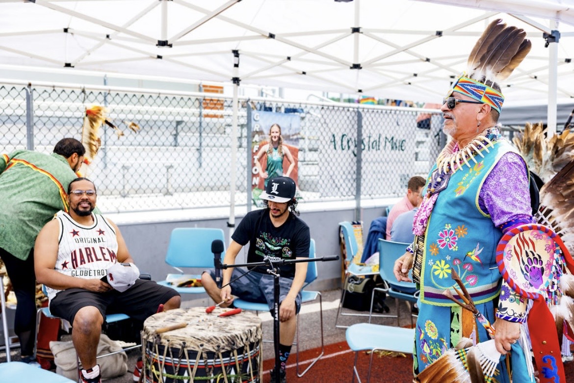 An elder in regalia standing beside the drum circle at the powwow