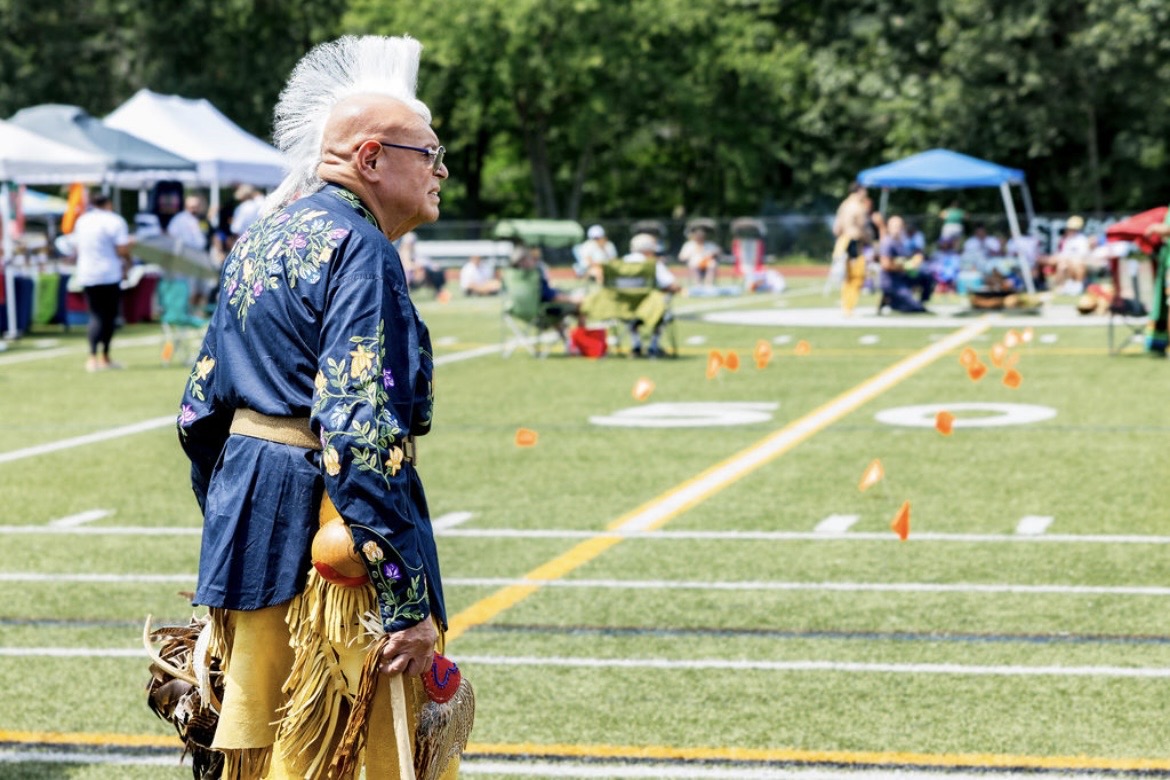 An elder in regalia with a roach headdress overlooking the powwow grounds