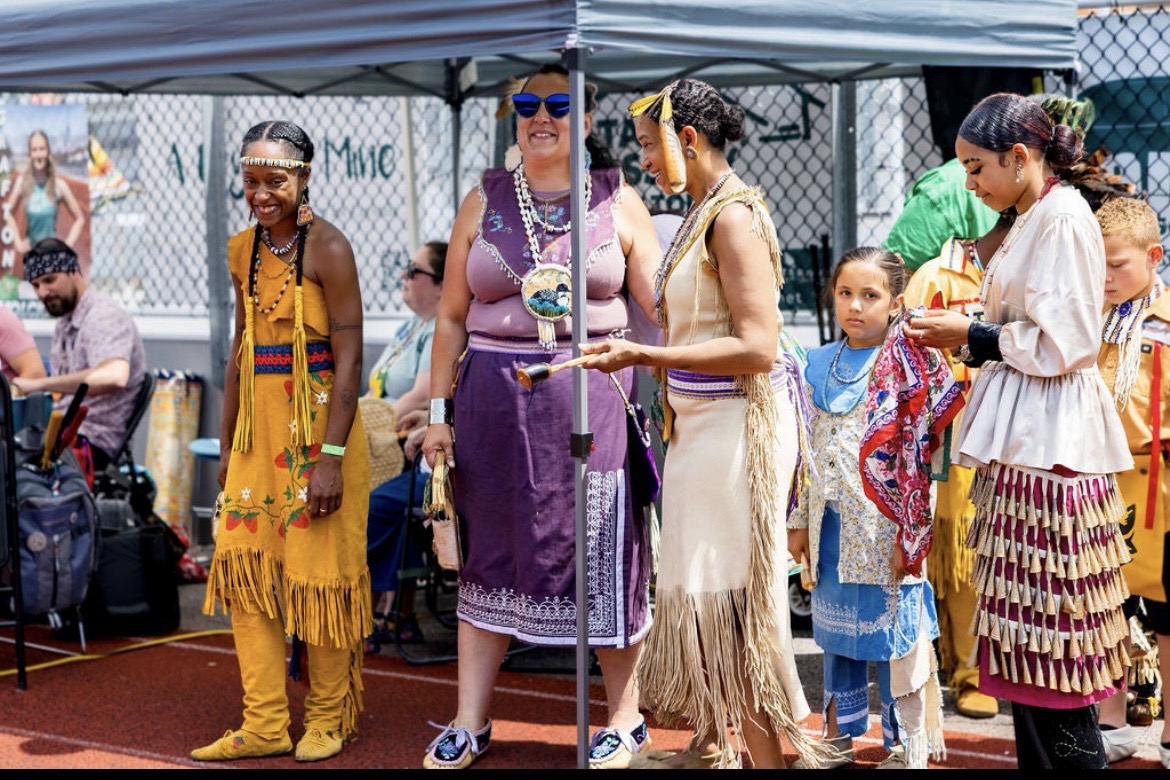Community members in regalia gathered together under a tent at the powwow