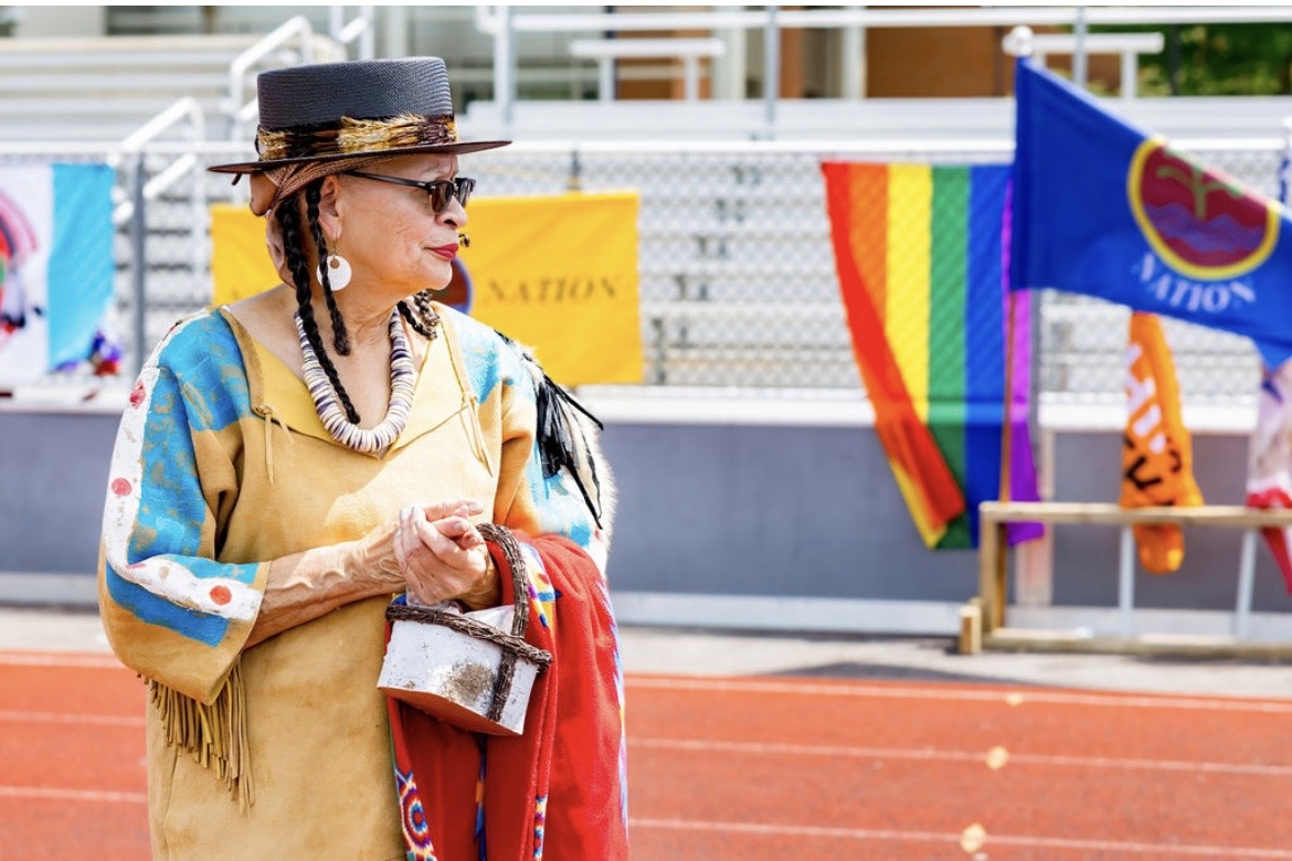 A woman in regalia carrying a basket near the flags at the powwow