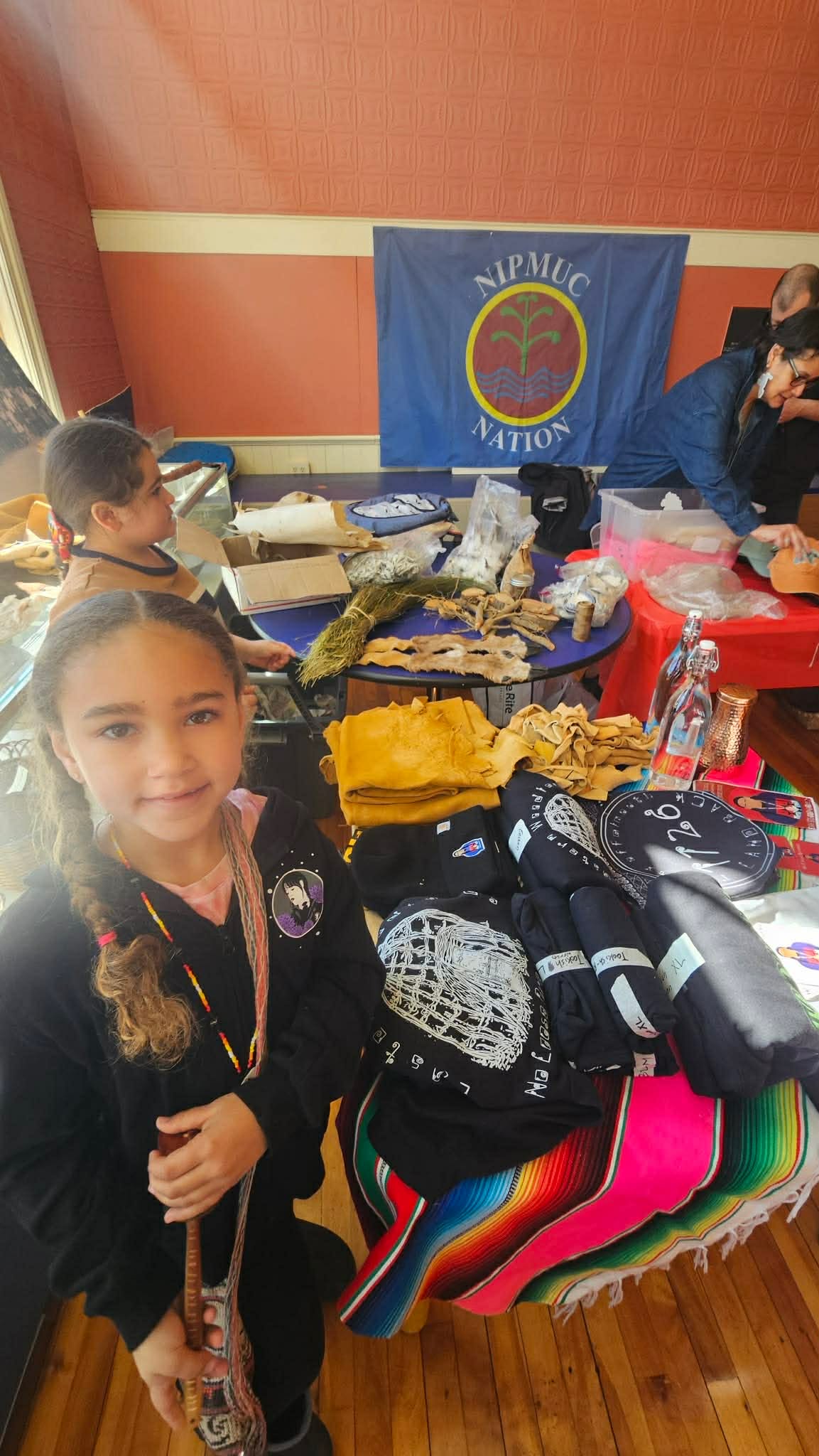 Two girls beside a table of Nipmuc Nation merchandise, blankets, and shirts