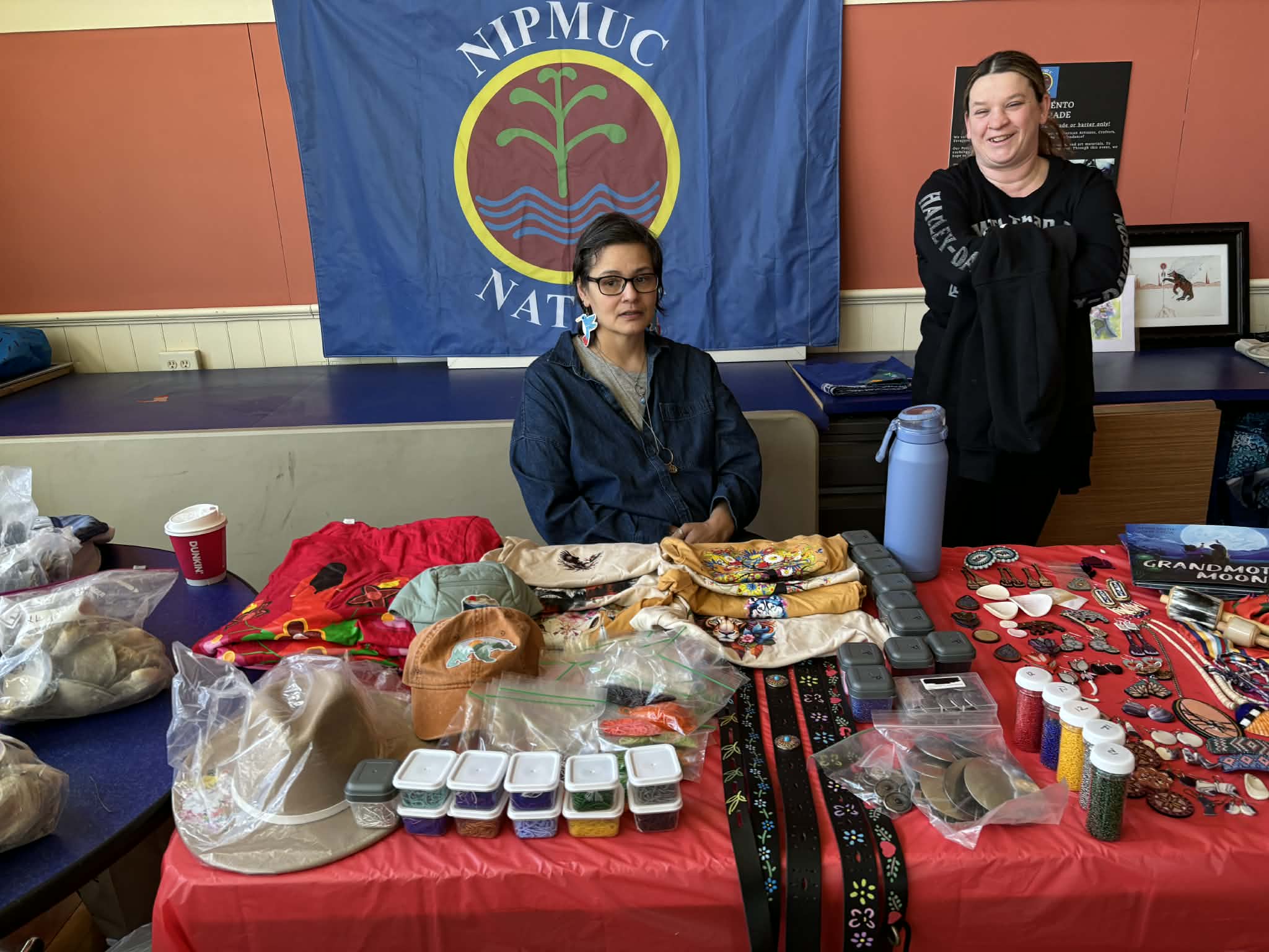 Two women at a vendor table of hats, jewelry, and craft supplies